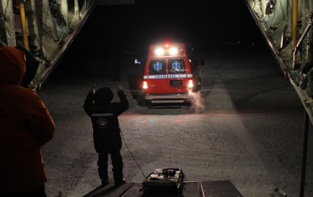 An ambulance moving towards the inside fo a military plane; an individual is directing the ambulance