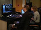 A man and woman view a brain scan on a monitor as MRI is scanning a patient in the next room.